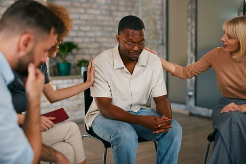 Vulnerable African American man receiving comforting support and touch from two women during an intimate multi-ethnic group therapy or counseling session indoors