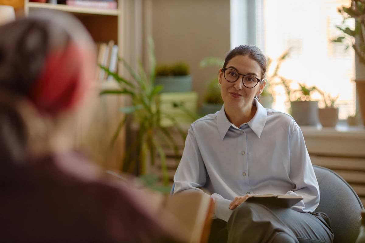 Caucasian young adult woman psychologist wearing glasses listening attentively to unseen patient during therapy session in office, holding clipboard, engaging in professional psychological counseling